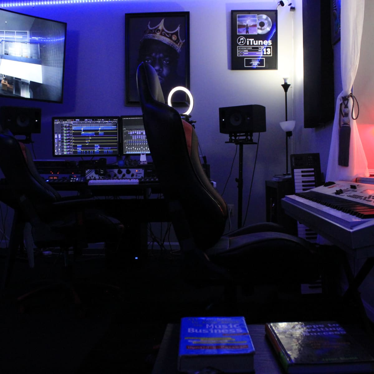 A dimly lit music studio features two black chairs facing a computer setup with multiple screens, speakers, and a keyboard, illuminated by ambient lighting.