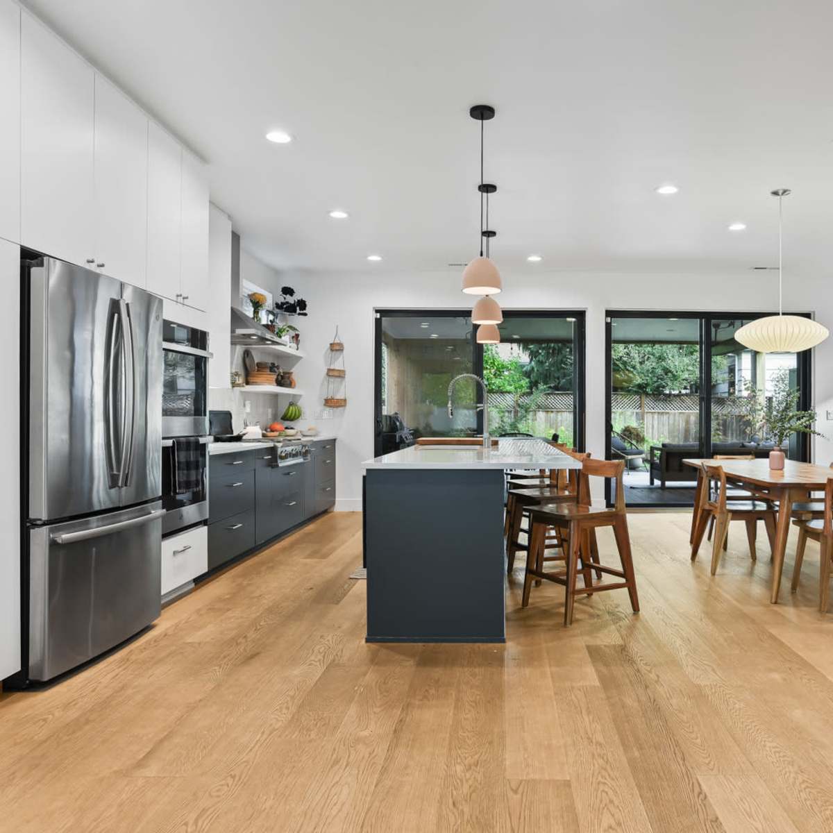 A modern kitchen features sleek cabinetry, stainless steel appliances, and a central island with seating, adjacent to a dining area surrounded by large windows.