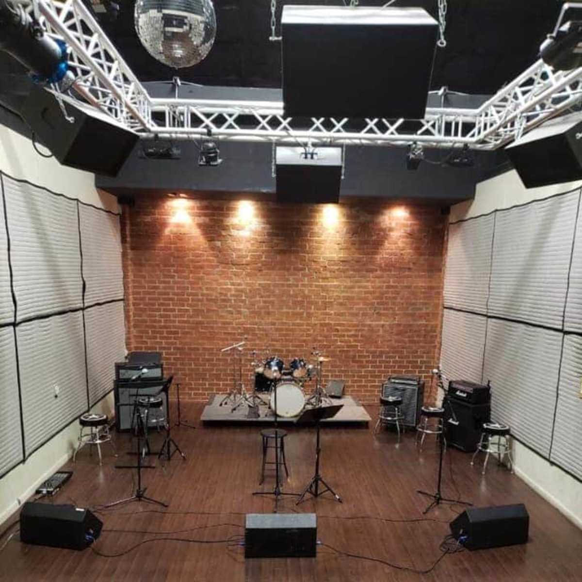 A music rehearsal space features a drum set on a small stage, surrounded by amplifiers and microphones against a backdrop of brick walls.