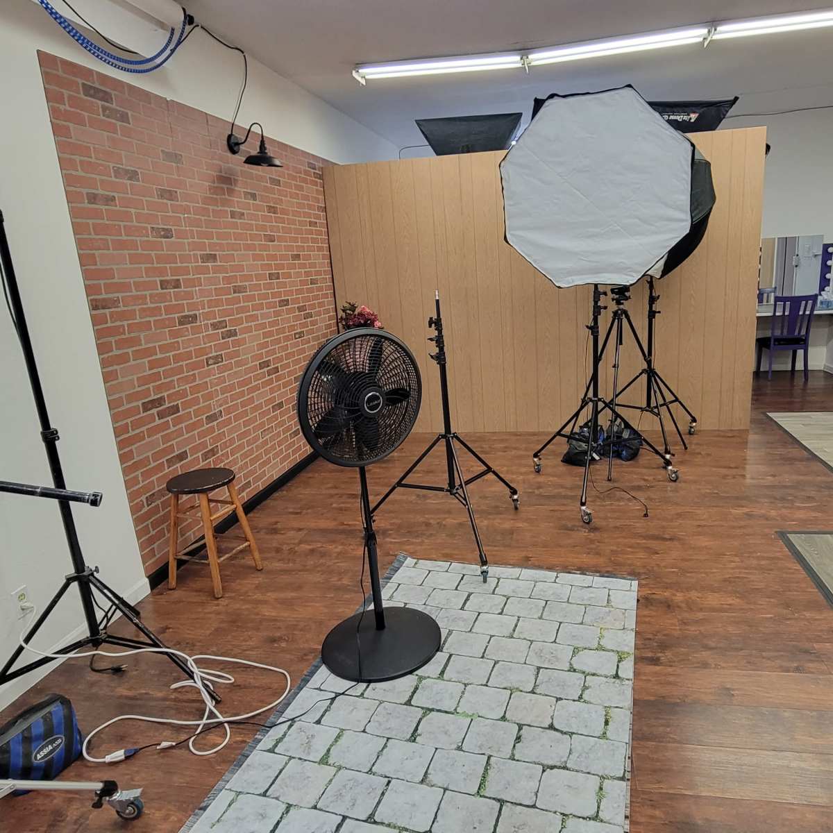 A photography studio with a brick wall backdrop, lighting equipment, a fan, and various types of furniture scattered around.