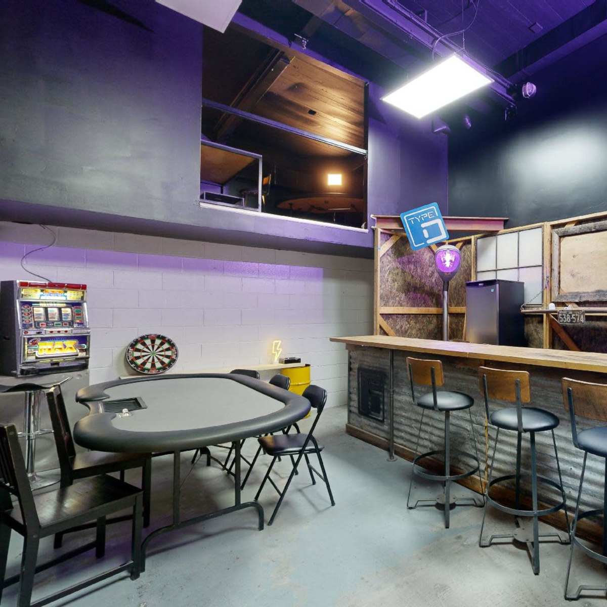 A small bar area with high stools, a poker table, and a dartboard, featuring industrial decor and purple lighting.