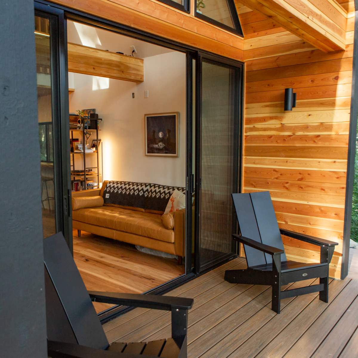 A cozy seating area inside a wooden cabin is visible through a large sliding glass door, framed by a covered deck with two black outdoor chairs.