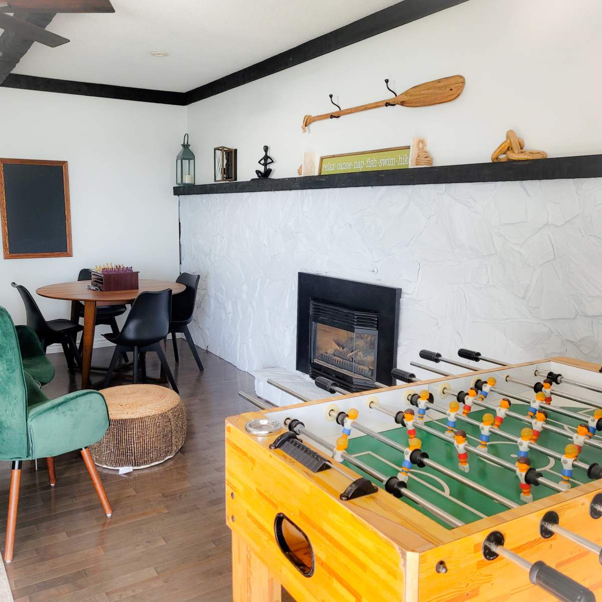 A cozy living area with a foosball table in the foreground, a round dining table, and a stone fireplace against a white wall.