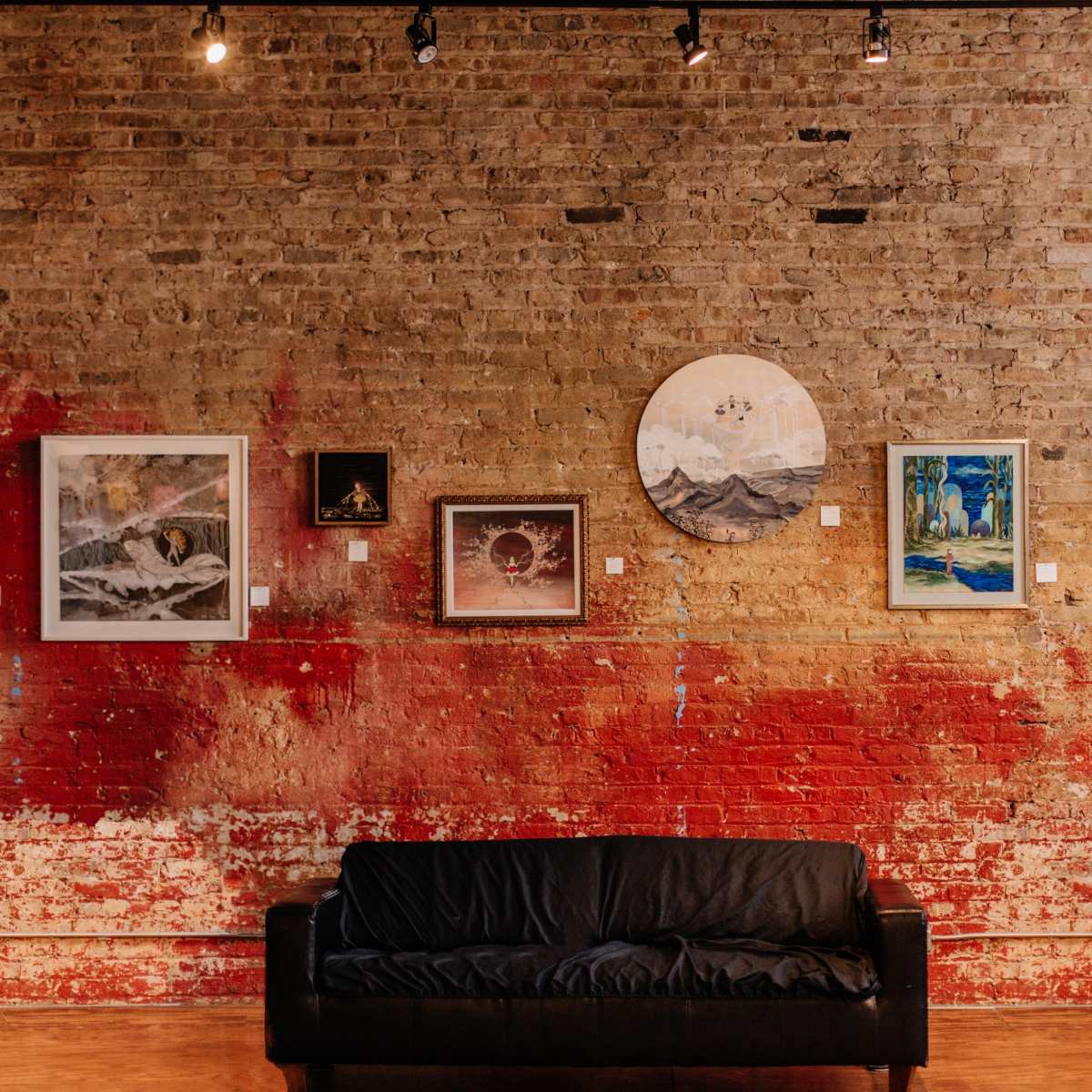 A gallery wall featuring various framed artworks displayed against a brick wall with a splash of red paint, alongside a black couch in the foreground.