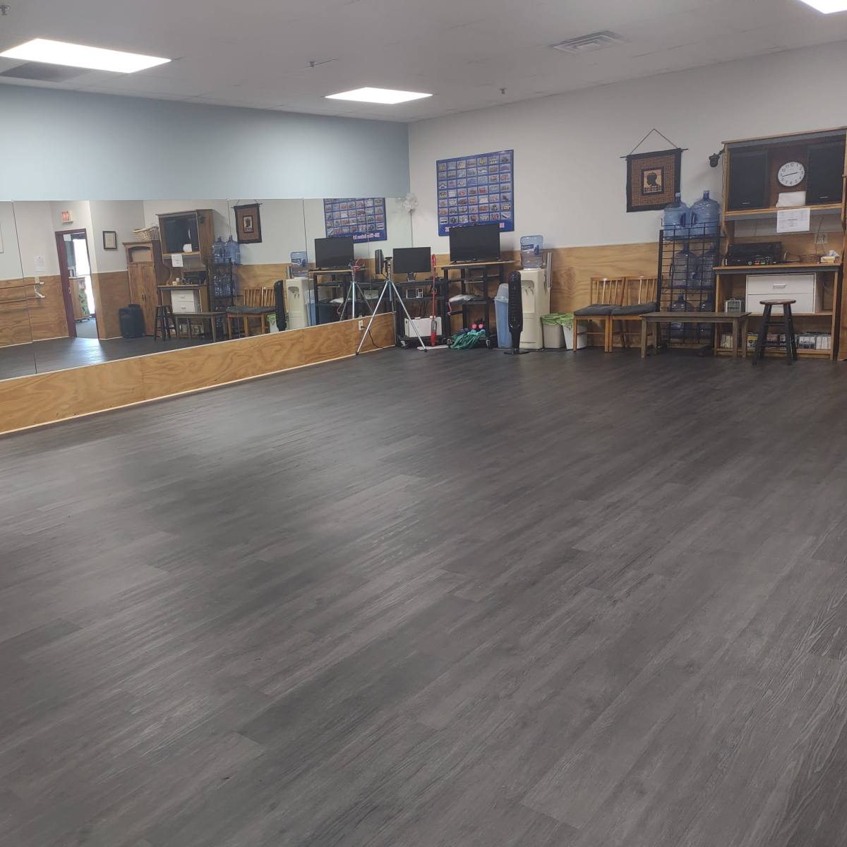 A spacious room with wooden flooring, a large mirrored wall, and various furniture pieces, including chairs and tables, along one side.