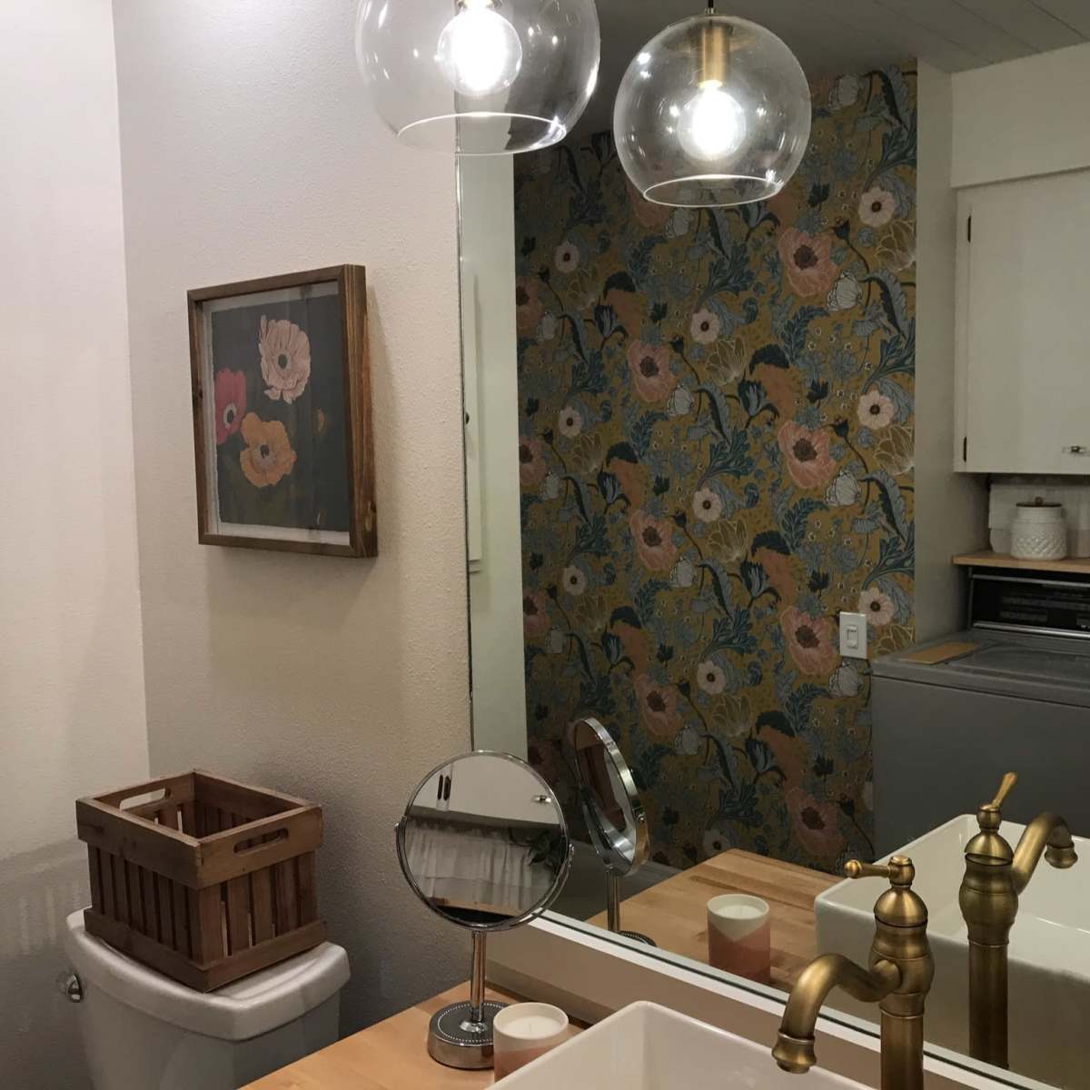 A bathroom features a wall with floral wallpaper, a large mirror, a sink with a gold faucet, a laundry machine, and a wooden basket for storage.