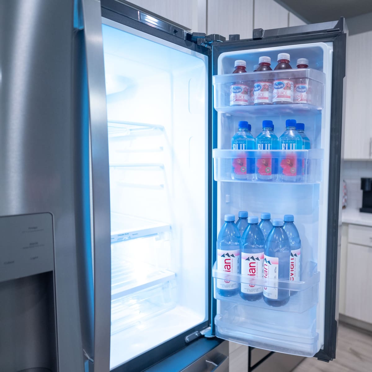 The interior of a refrigerator with shelves containing bottles of water and flavored drinks, alongside a bright kitchen backdrop.