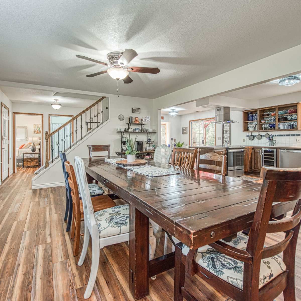 A dining area with a large wooden table surrounded by chairs, adjacent to a living space and kitchen with a staircase in the background.