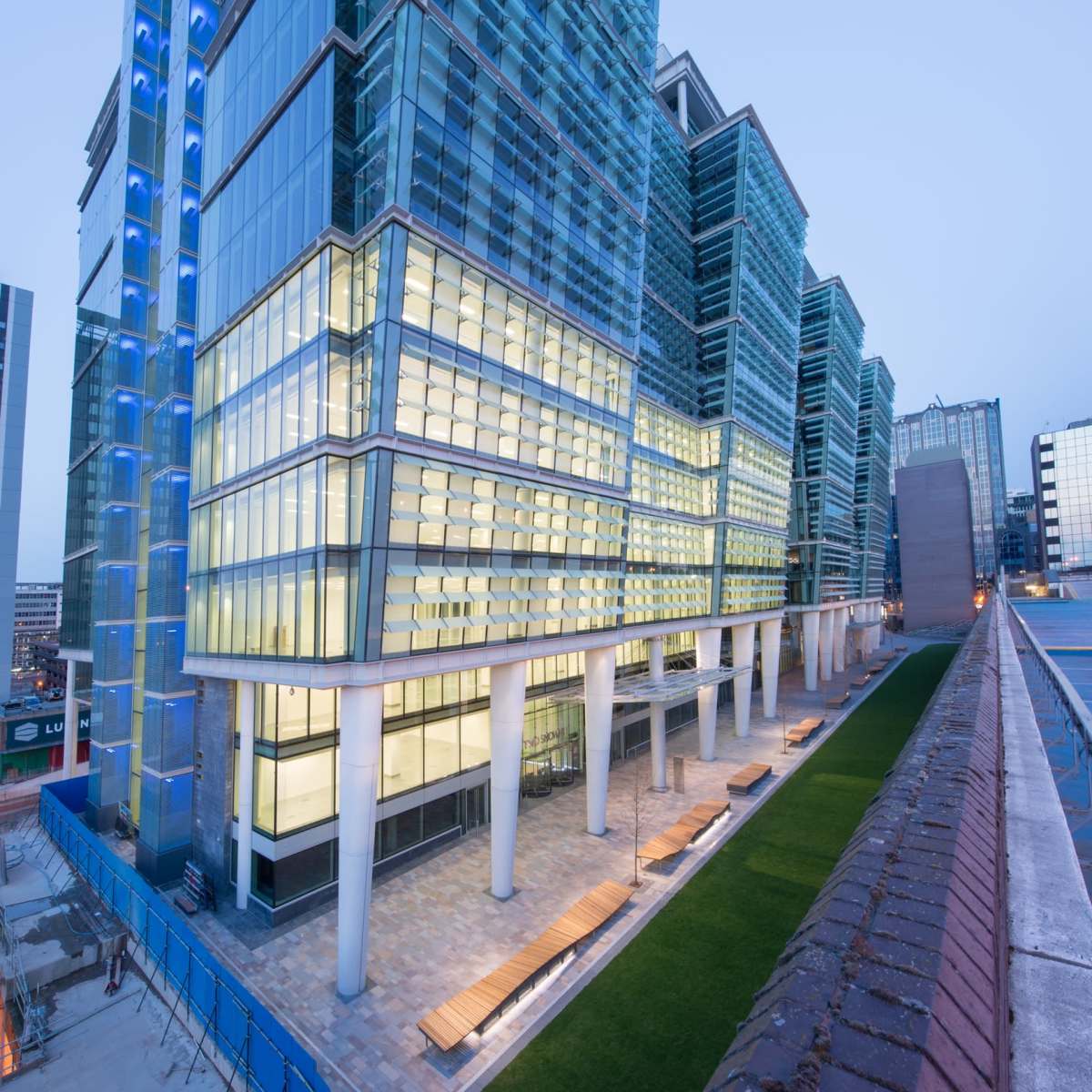 A modern multi-story glass building with a wide entrance, surrounded by a landscaped area and city skyline in the background during twilight.