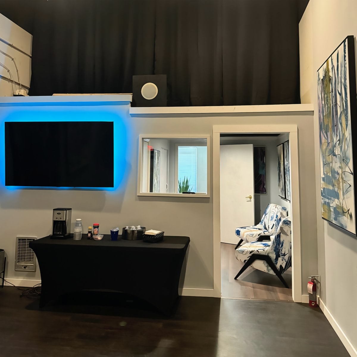 A modern room with a black table displaying drinks, a large TV on the wall with blue lighting, and a doorway leading to another area featuring a patterned chair and artwork.