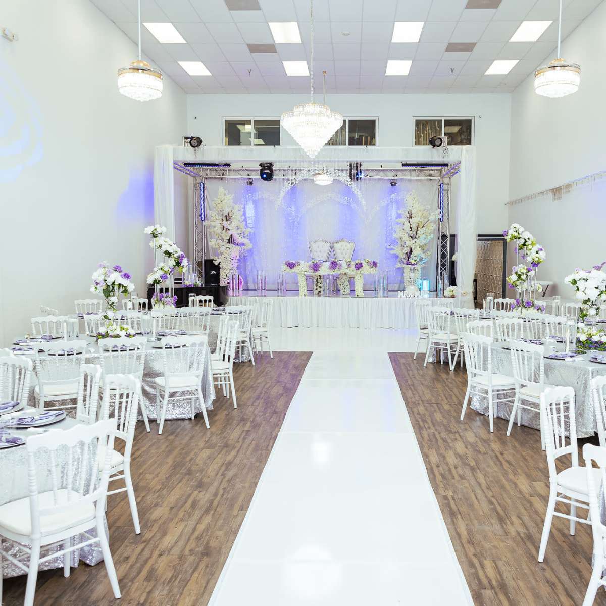 A spacious banquet hall set up for an event, featuring round tables with white tablecloths and centerpieces, and a stage at the front adorned with floral arrangements and decorative lighting.