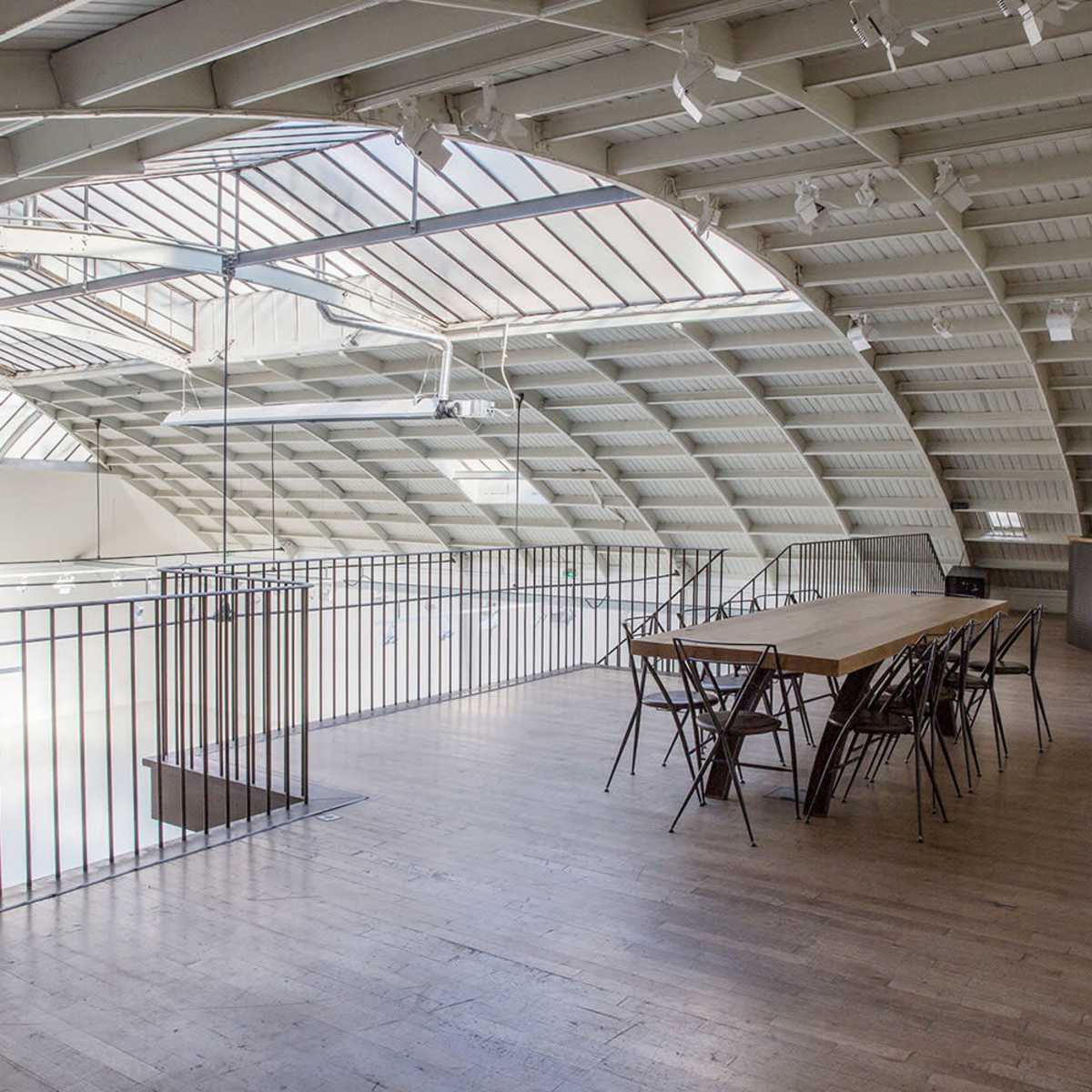 A modern, open loft space with a long wooden table and chairs, featuring a curved roof and large skylights.