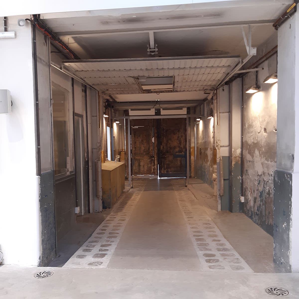 A long, narrow corridor leading to a pair of wooden doors, with walls that have peeling paint and exposed concrete flooring.