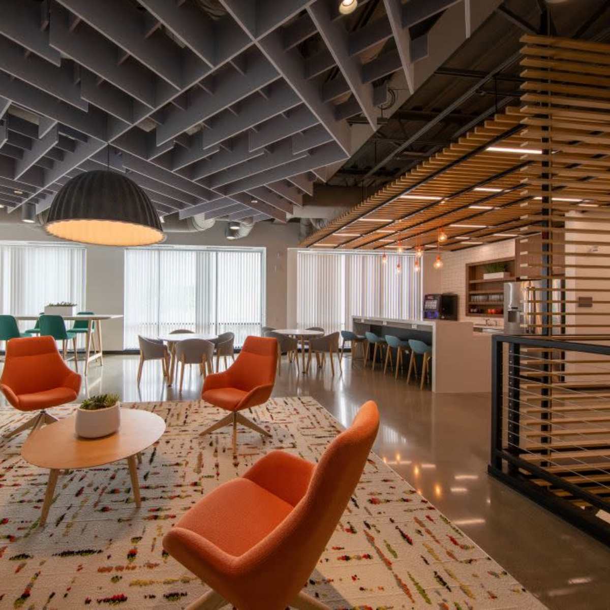 A modern office lounge area with orange chairs, a round wooden table, and large windows letting in natural light, complemented by a patterned rug and a kitchenette in the background.