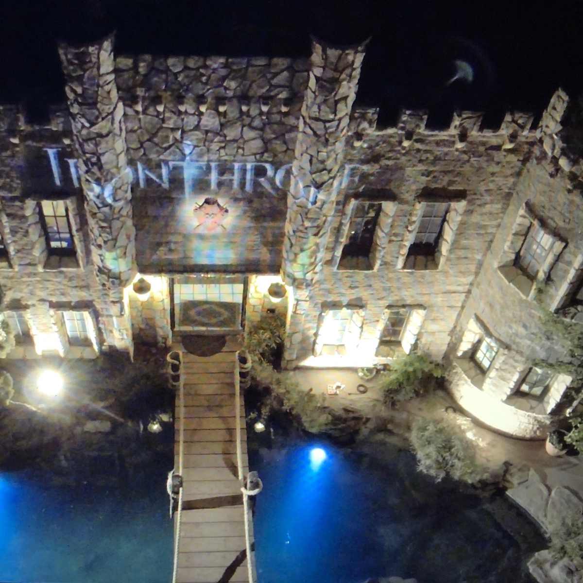 A stone mansion with a bridge over a water feature is illuminated at night, featuring a projection on its facade.