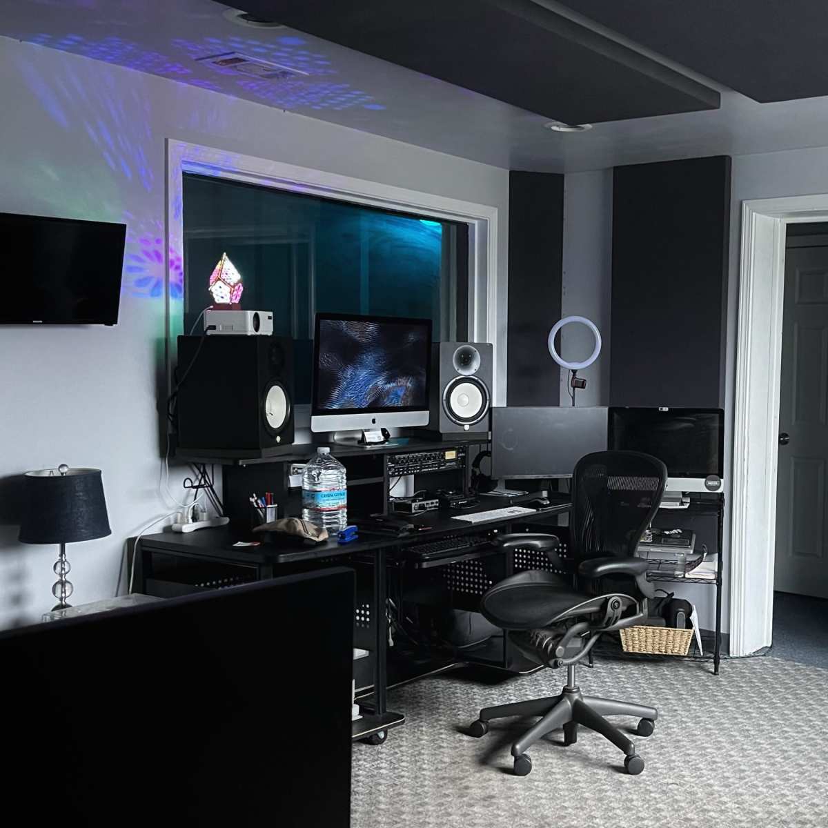 A music production studio featuring a desk with multiple computers and audio equipment, a large window, and a black couch.