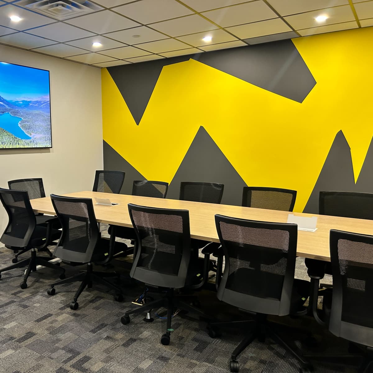 A modern conference room with a long table, black office chairs, and a large screen displaying a landscape photo on a yellow and gray geometric wall.