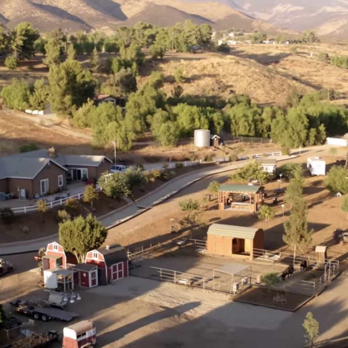 A rural landscape featuring a cluster of houses, a barn, and several paddocks surrounded by hills and trees.