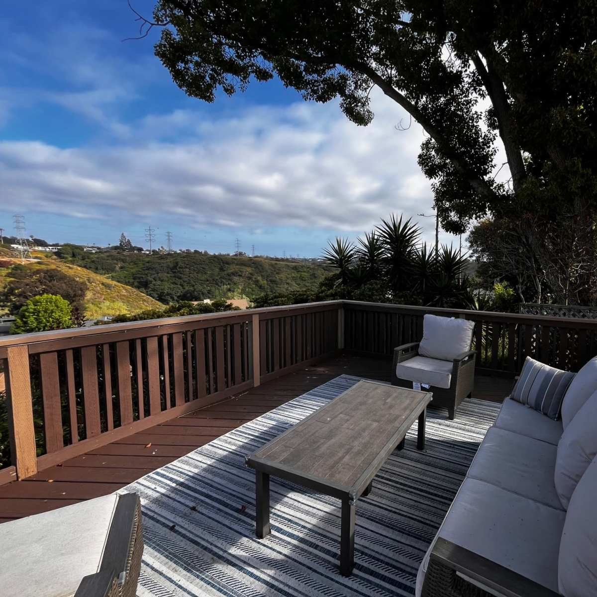 A wooden deck with outdoor furniture, including a sofa and a coffee table, overlooking a hilly landscape under a partly cloudy sky.