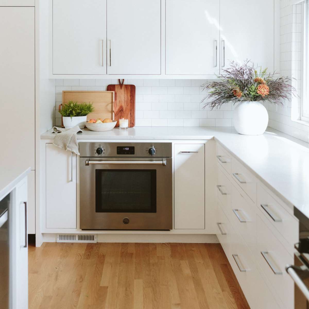 A modern kitchen features white cabinetry, a stainless steel oven, and a countertop with a cutting board and a bowl of fruit.