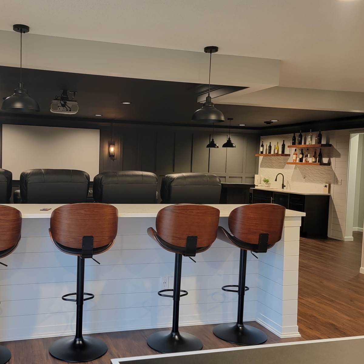 A modern home theater with five high-backed bar stools arranged at a white counter, facing a dark wall with a projector screen and leather seating in the background.