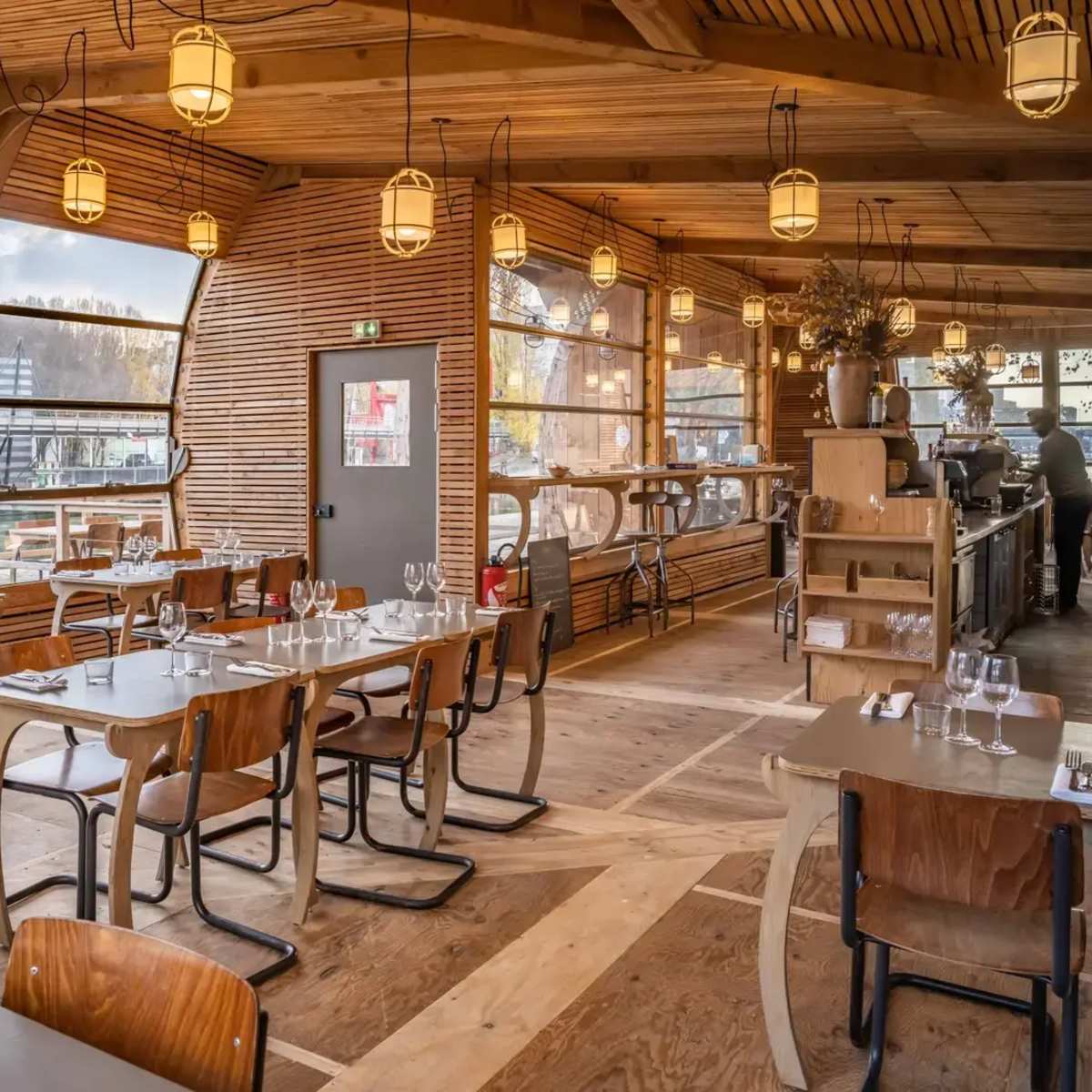 A modern restaurant with wooden interiors, featuring tables and chairs arranged near large windows overlooking a waterfront.