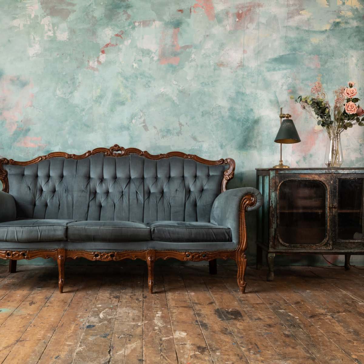 A vintage teal velvet sofa is centered in front of a textured, colorful wall, accompanied by a potted plant, a small lamp, and a glass cabinet.