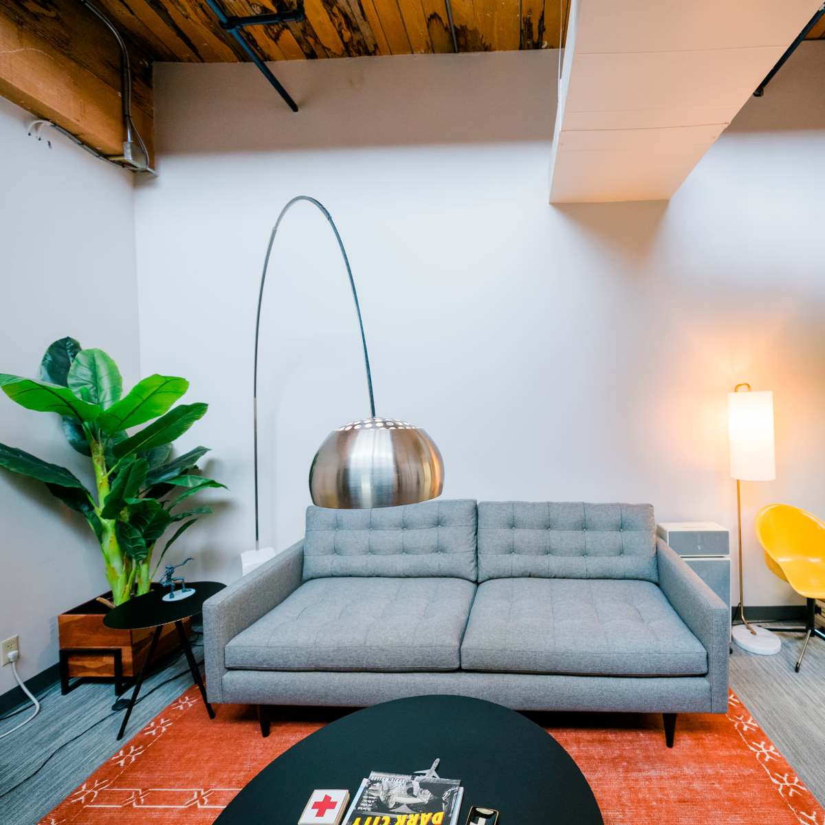 A modern office lounge features a gray sofa, an arc floor lamp, a tall indoor plant, and a circular coffee table on a patterned rug.
