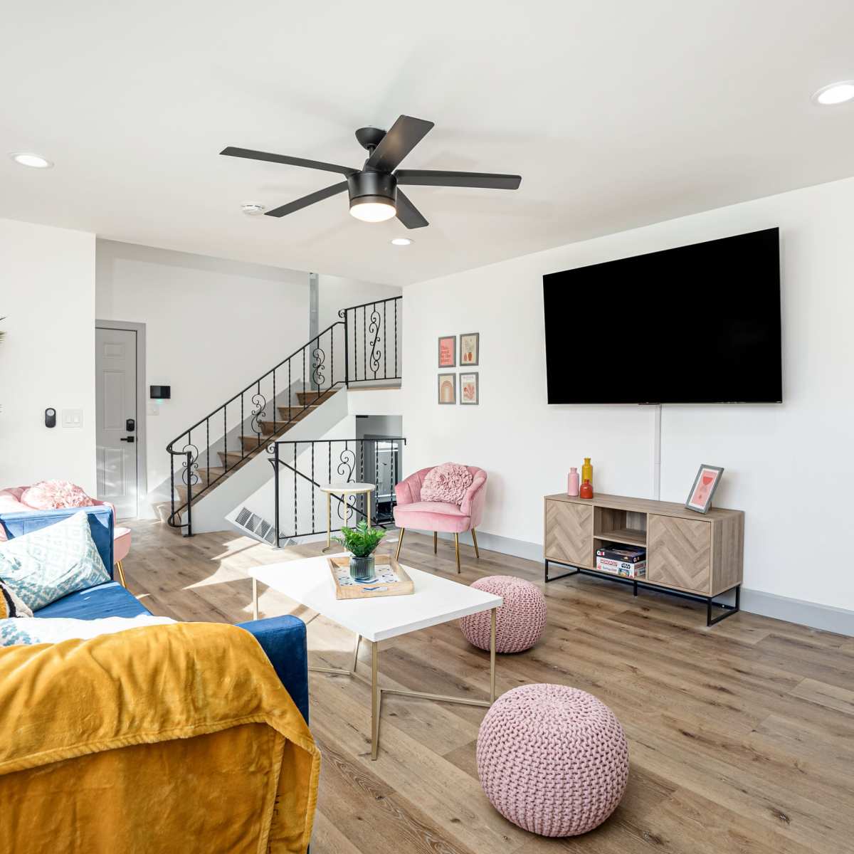 A modern living room featuring a blue sofa, pink armchairs, a wooden TV stand, and a staircase in the background.