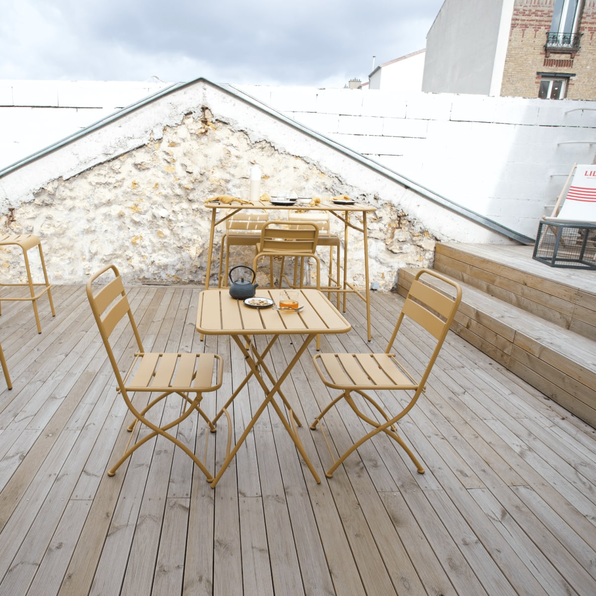 A terrace features yellow metal furniture arranged on wooden decking next to a white wall.