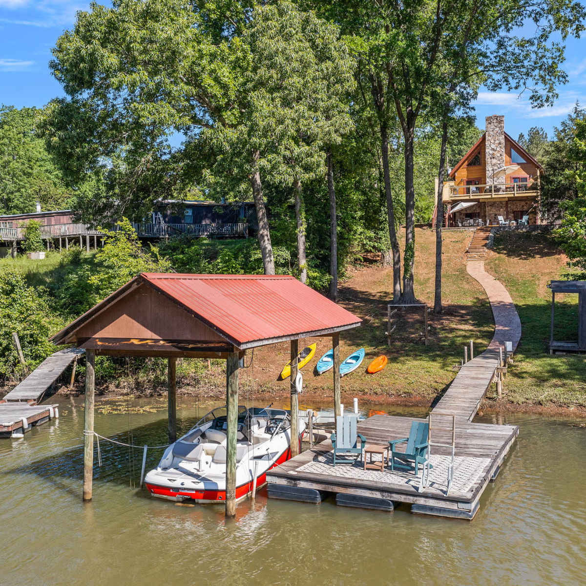 A boat dock with a red-roofed shelter is located on a calm waterway, surrounded by trees and a pathway leading to a house on a hillside.
