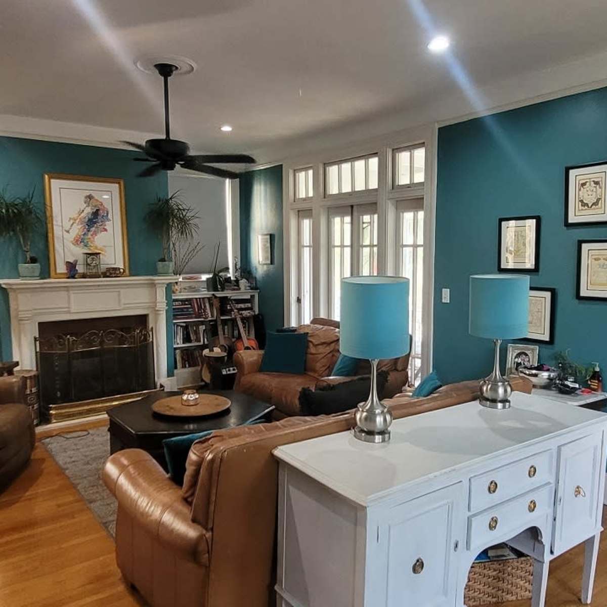A living room with teal walls, a fireplace framed by bookshelves, and two brown armchairs opposite a white console table with blue lamps.