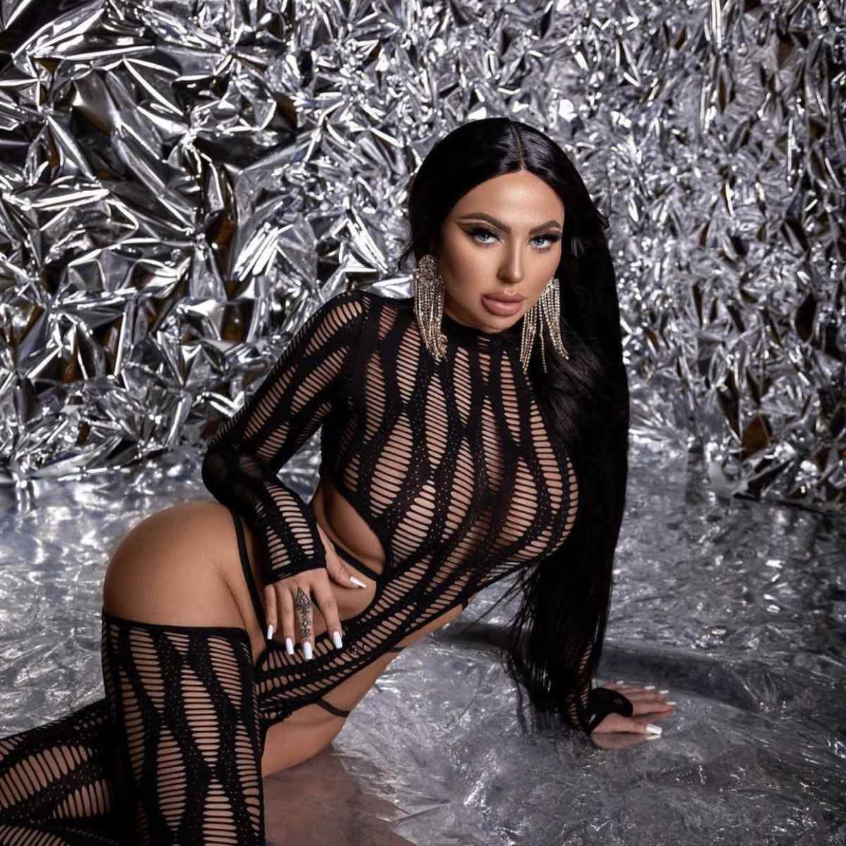 A person poses on a reflective surface in a black mesh outfit against a shiny, crinkled metallic backdrop.