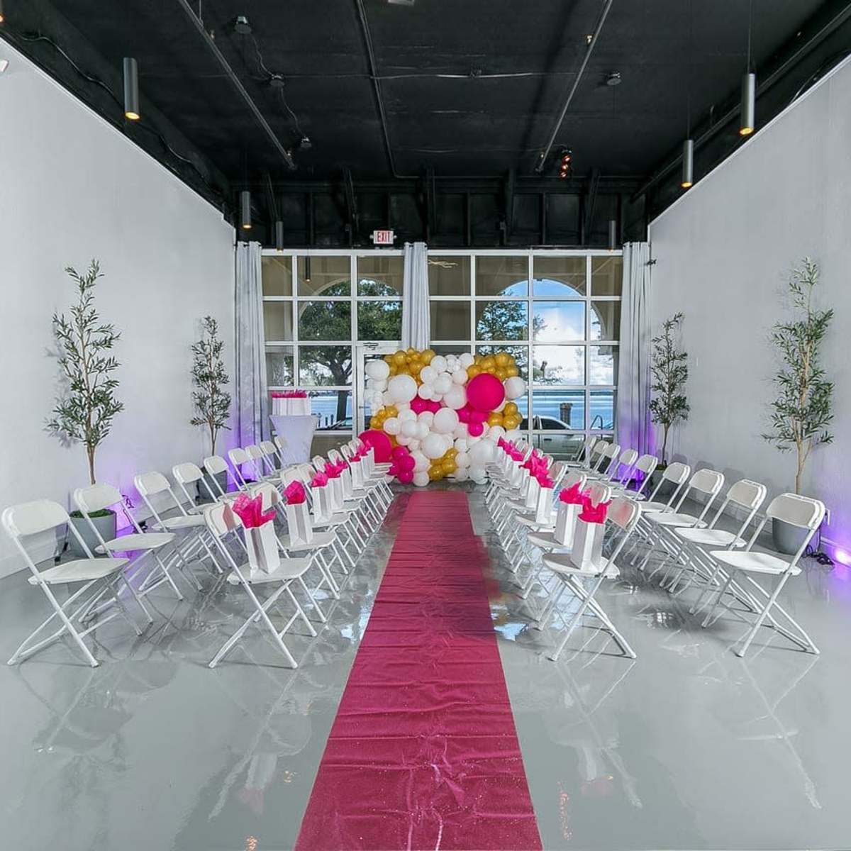 A ceremony space features rows of white chairs along a red carpet, with a backdrop of colorful balloons and potted trees.