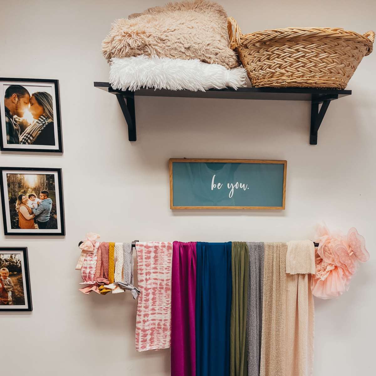 A wall with framed photos on the left, a shelf holding a basket and a cozy blanket on top, and a display of colorful fabrics hanging below.