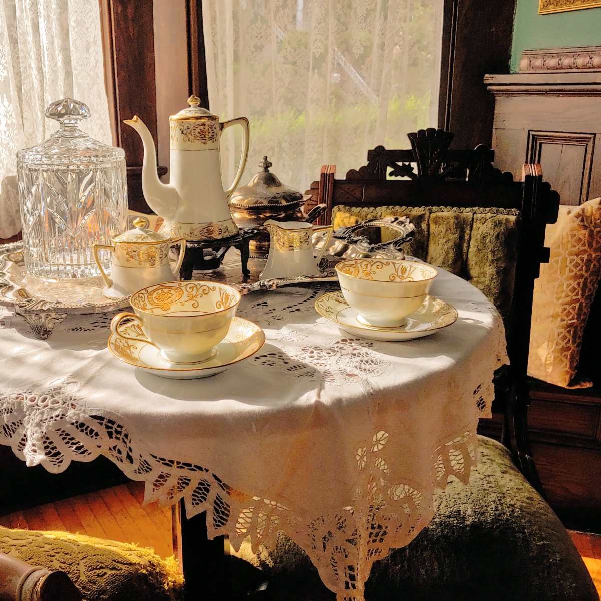 A polished tea set with ornate designs is arranged on a lace tablecloth beside a doll in a vintage room with sunlight streaming through a window.