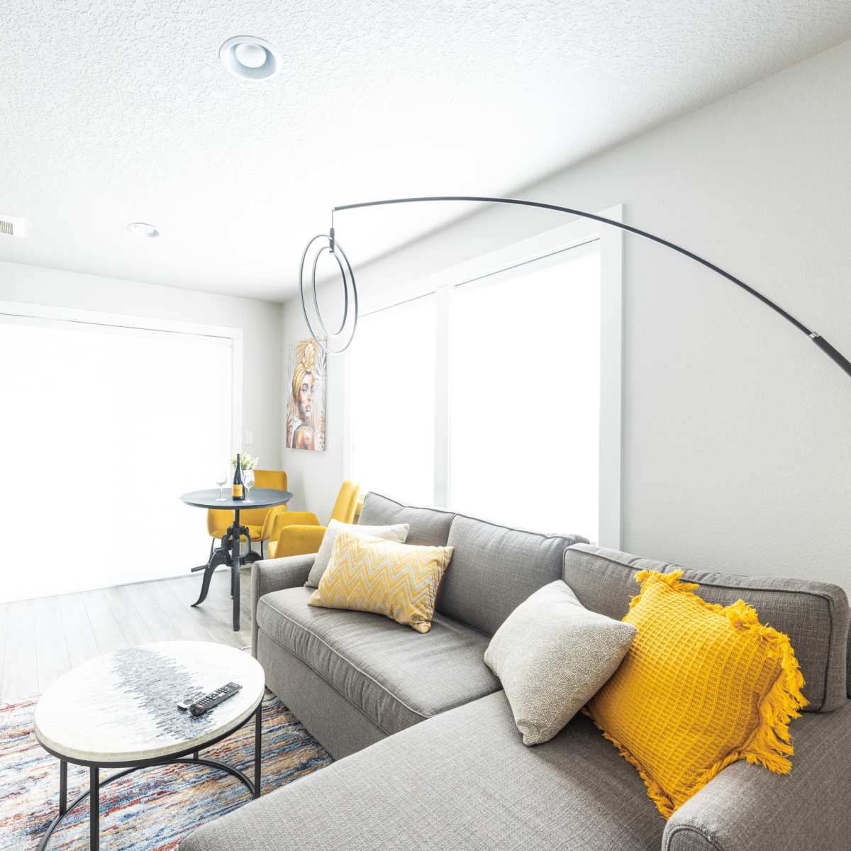 A modern living room with a gray sofa, yellow throw pillows, a circular coffee table, and large windows allowing natural light to fill the space.