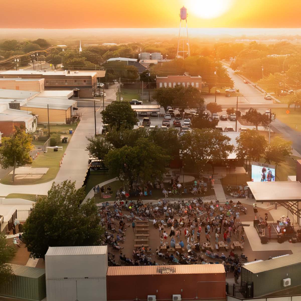A large outdoor gathering at sunset in a town center, with people seated in a park area facing a screen amidst surrounding buildings.