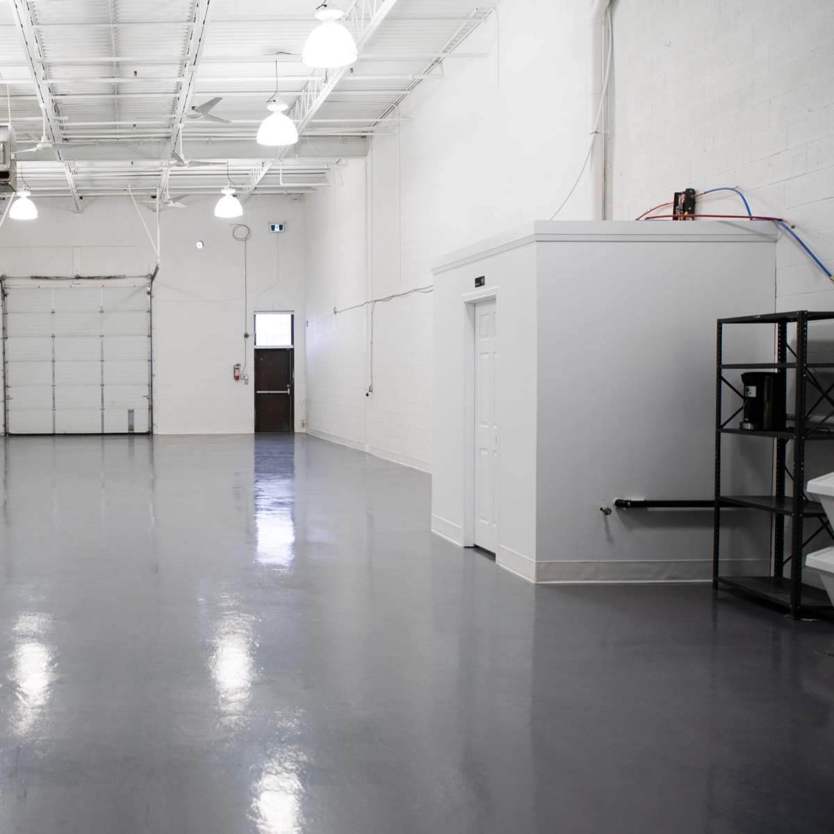 A large, empty industrial space with glossy gray flooring, white walls, and a roll-up garage door at one end.