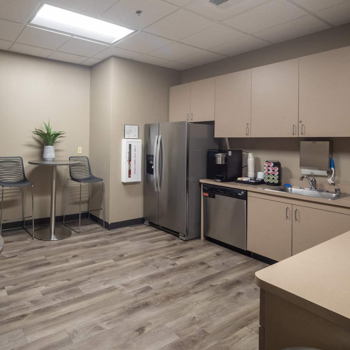 A modern break room with a kitchenette, including a refrigerator, coffee maker, and two high tables.