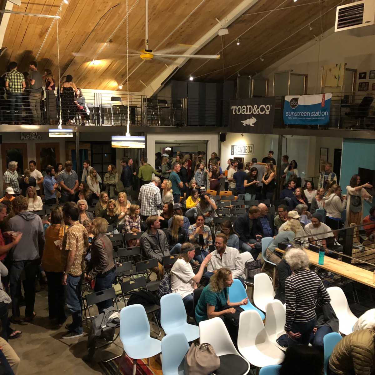A crowded venue filled with people engaging in conversation, seated and standing, with wooden beams and banners visible in the background.