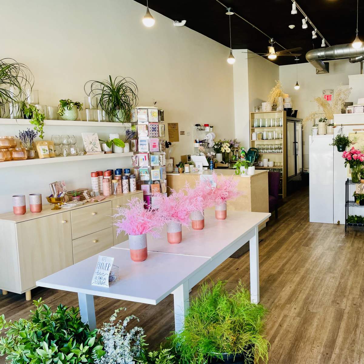 A neatly arranged floral shop features shelves of plants and floral arrangements, with a central table displaying pink decorations and a clean, organized layout.