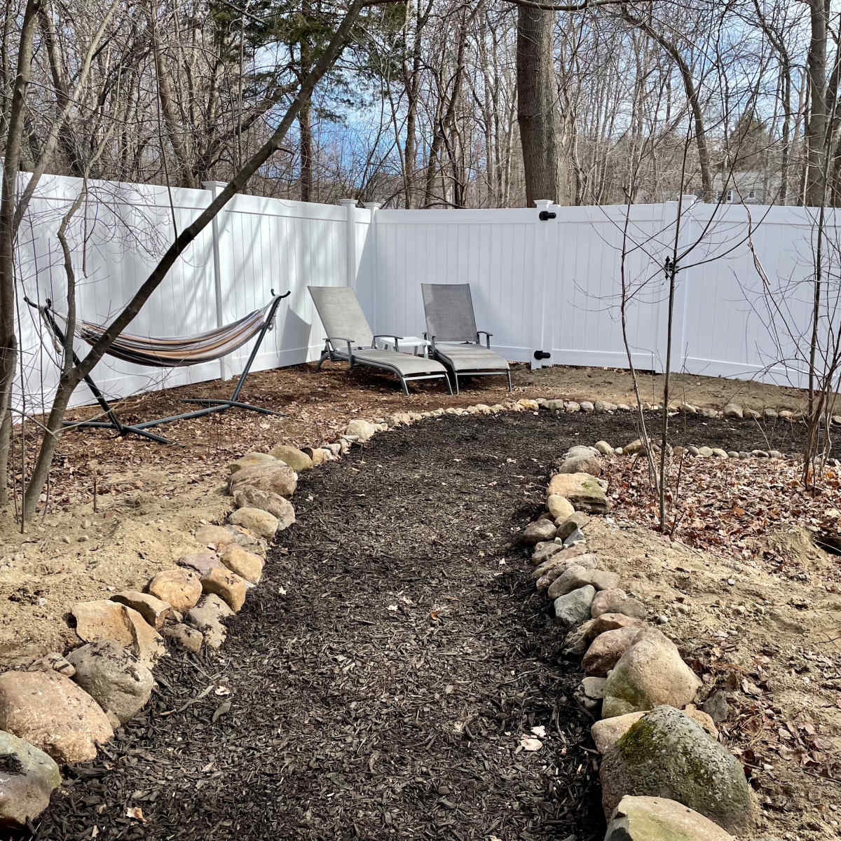 A garden area with a gravel path bordered by stones, two lounge chairs, a hammock, and a white fence in a leafless environment.