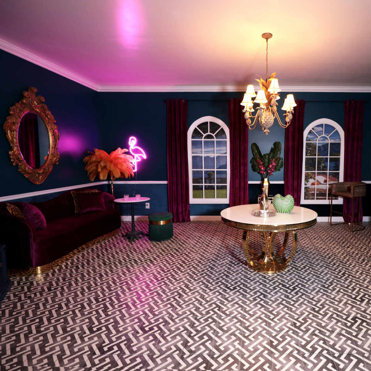 The interior of the room features a dark blue wall with a decorative chandelier, a round table with a gold base, a velvet couch, and large windows.