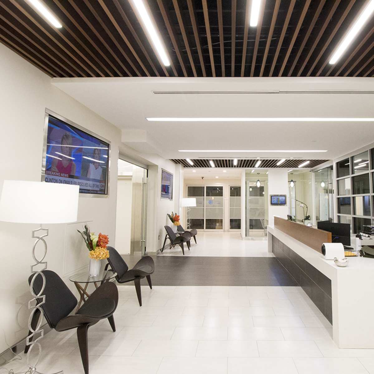 A modern office reception area with a front desk, several chairs, large windows, and two television screens mounted on the walls.