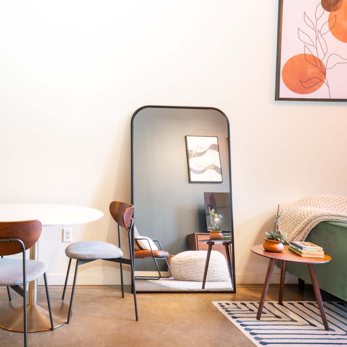 A small dining area featuring a round white table surrounded by four wooden chairs, with a large mirror and a framed artwork on the wall.
