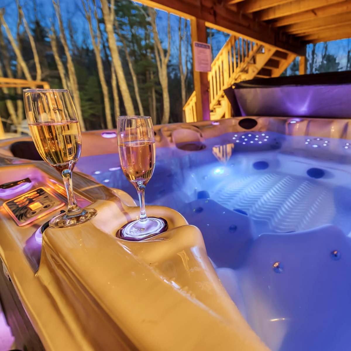 A hot tub under a wooden gazebo, with two champagne glasses placed on its edge and colorful lights illuminating the water.