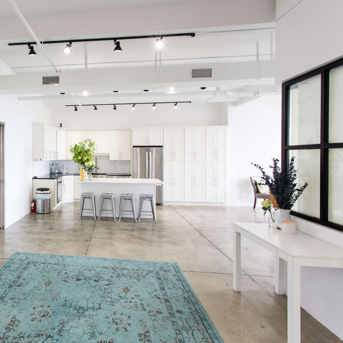A bright, modern kitchen and dining area with a concrete floor, white cabinetry, and a turquoise area rug.