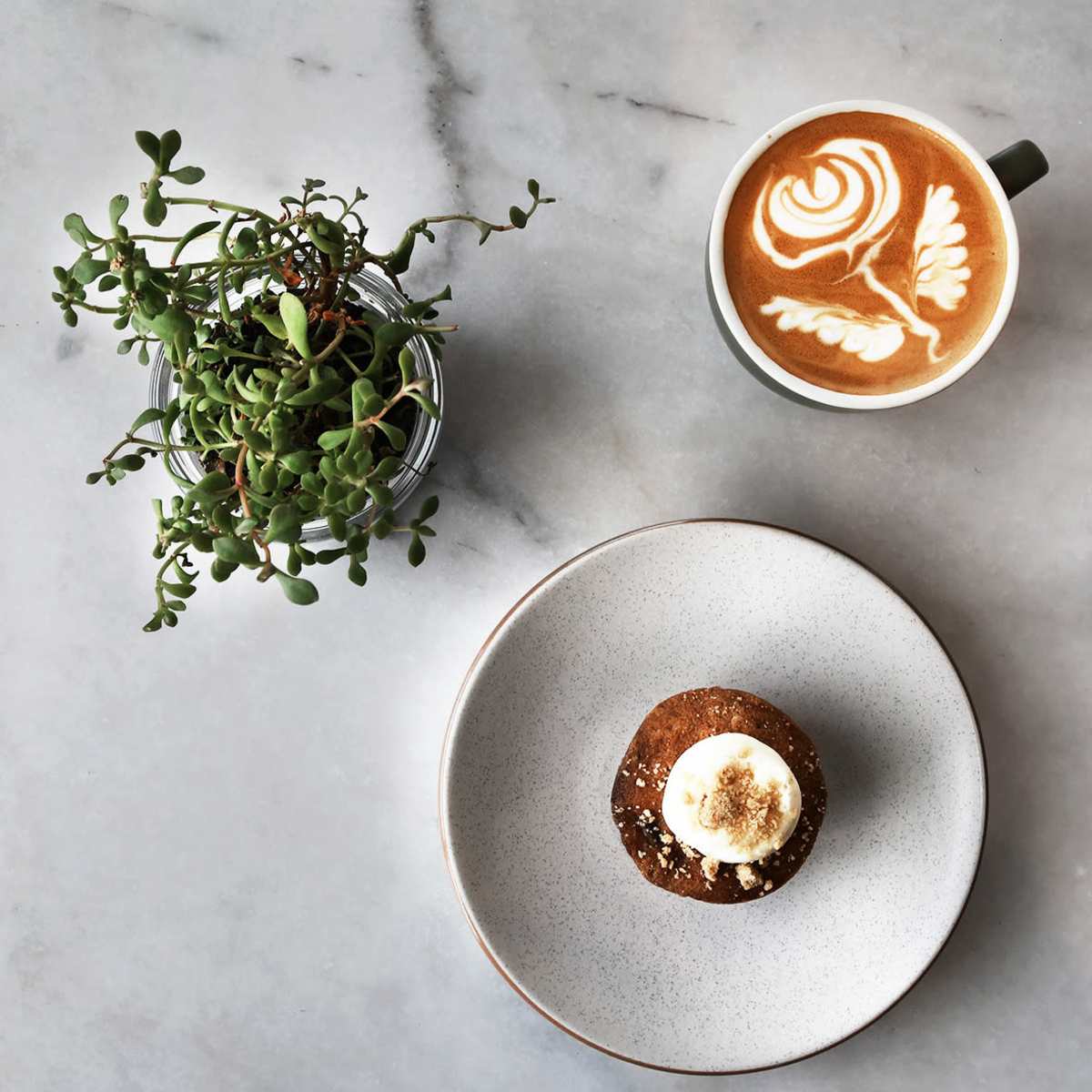 A muffin topped with frosting sits on a plate next to a cup of latte with a floral design, accompanied by a small potted plant on a marble surface.