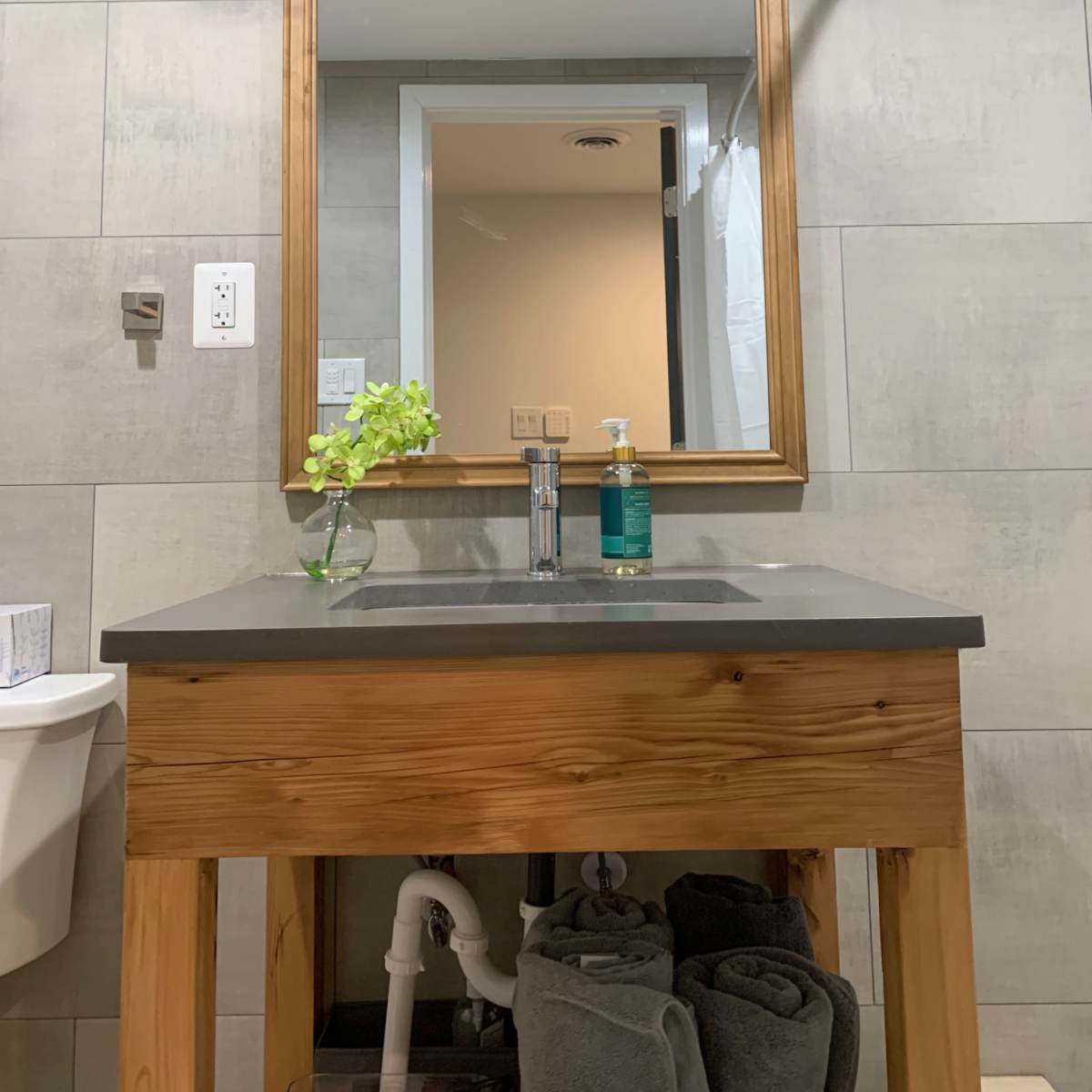 A wooden vanity with a gray countertop is positioned in front of a large mirror reflecting a doorway, with a small plant and liquid soap on the countertop.